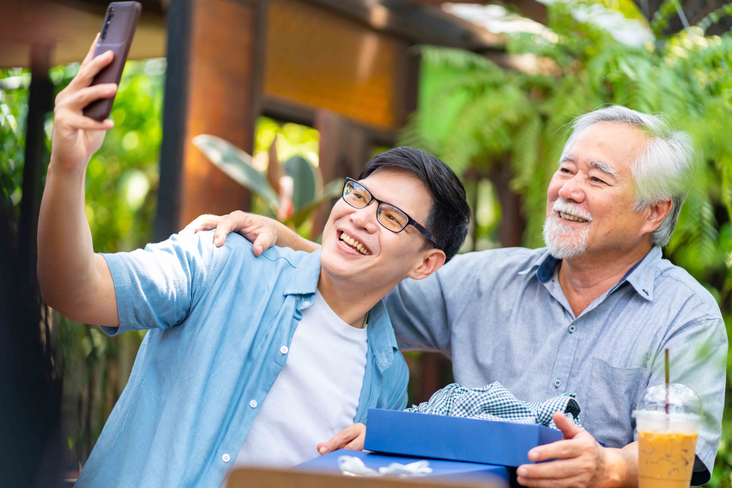 A young man takes a selfie of him and an older man outside at a table.