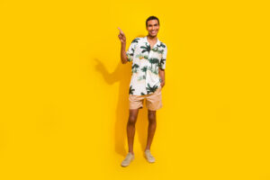 Full length photo of nice young male pointing empty space promo wear trendy palms print garment isolated on yellow color background.