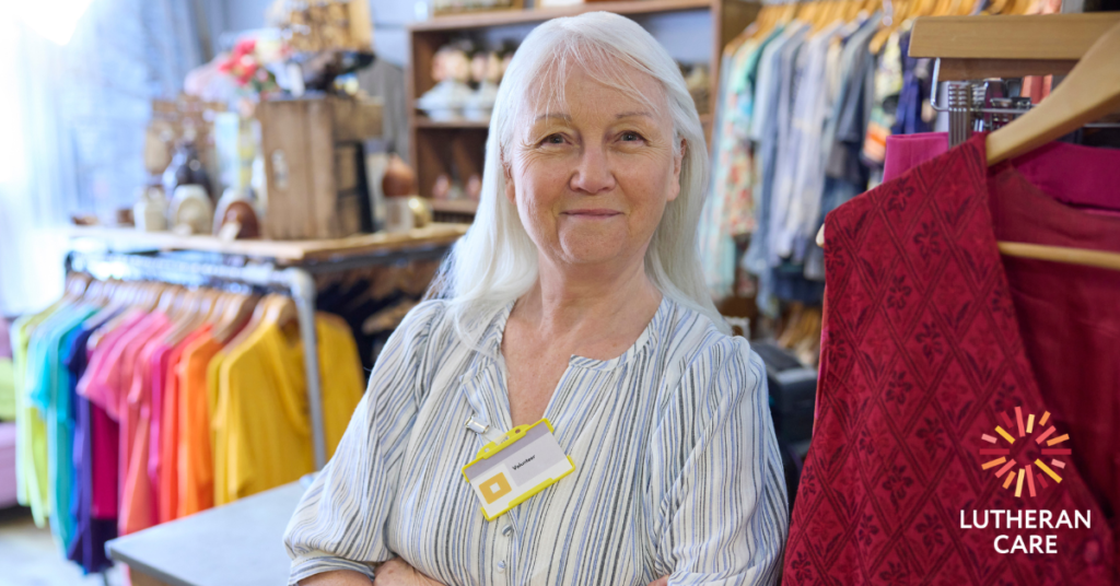 A lady with white hair and a light shirt is standing in an op shop among bright coloured clothes. The Lutheran Care logo appears in the bottom right hand corner.